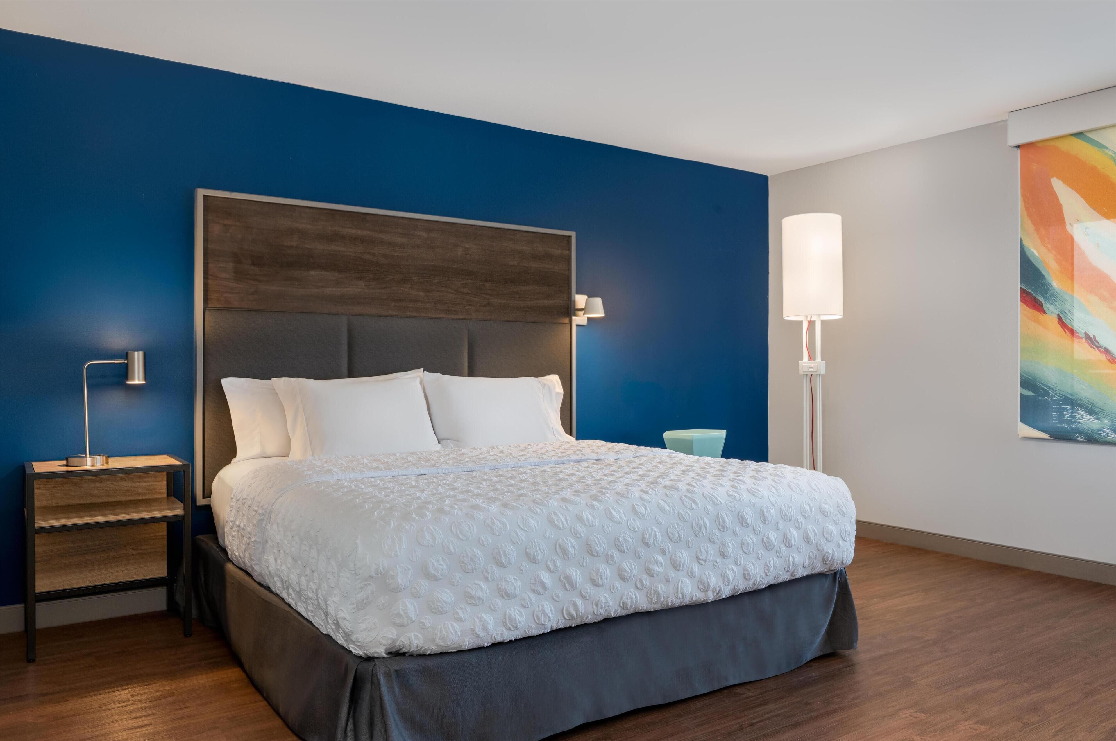 A modern hotel room with a large bed, a nightstand, a bedside lamp, and a colorful abstract painting on the wall.