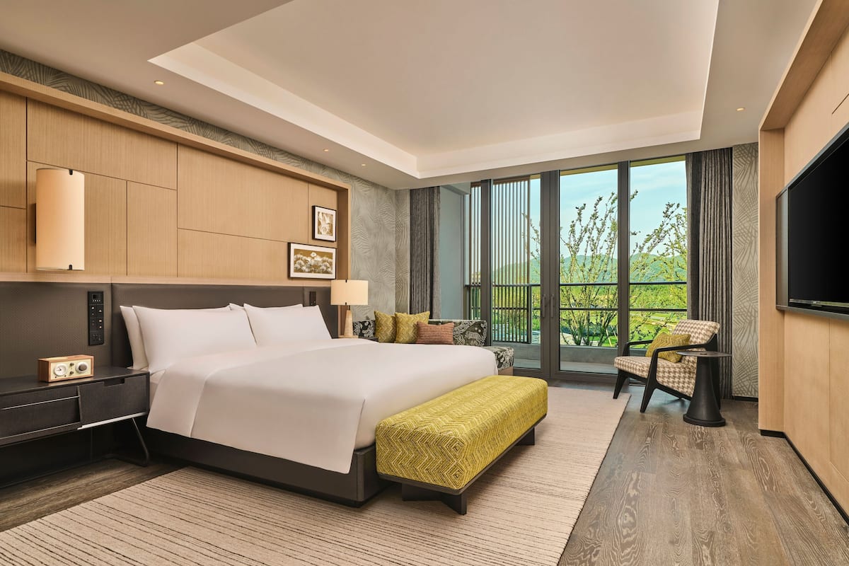 A modern hotel room with a large bed, a TV, a desk, and a balcony with a view of greenery.