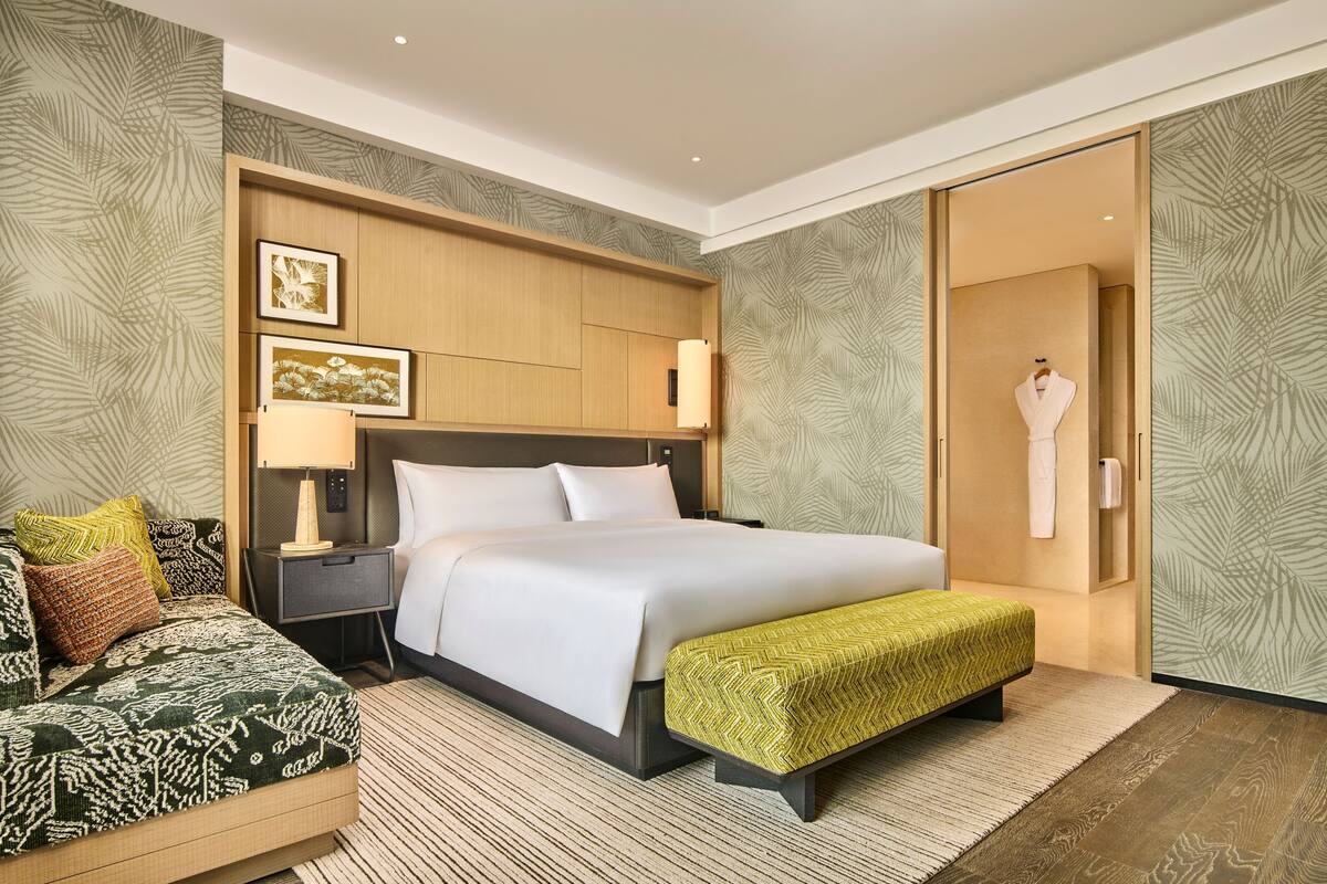 A hotel room with a large bed, a bedside table with a lamp, a bench, and a wardrobe.