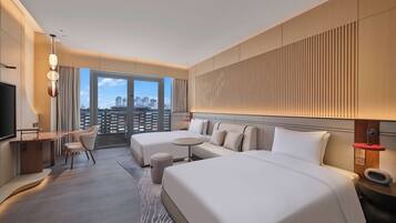Habitación de hotel moderna con una cama grande, un escritorio y una silla. Se aprecia una vista de la ciudad a través de amplias ventanas.
