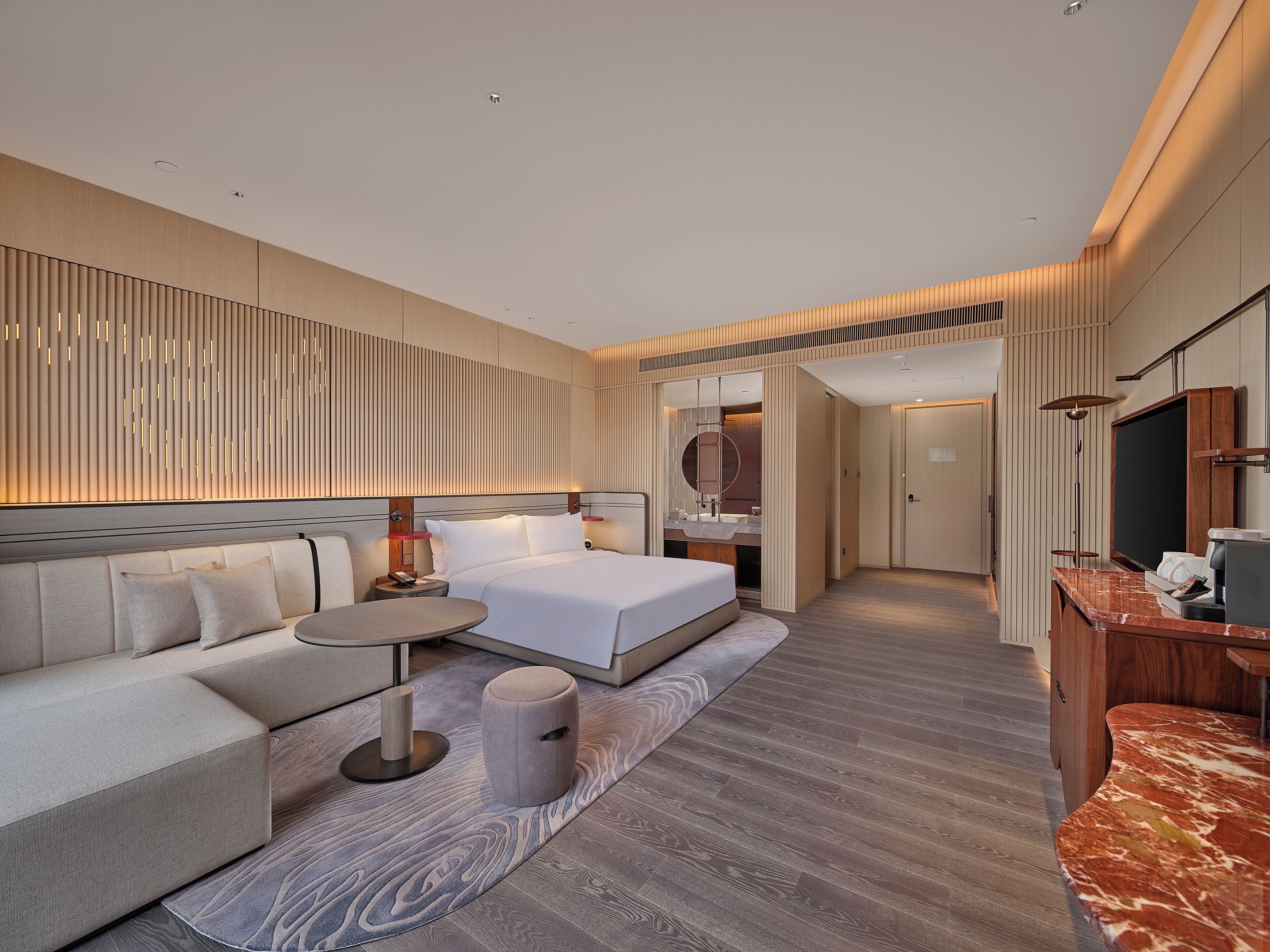 A modern hotel room with a sofa, a round table, a bed, a mirror, a TV, and a wooden floor.