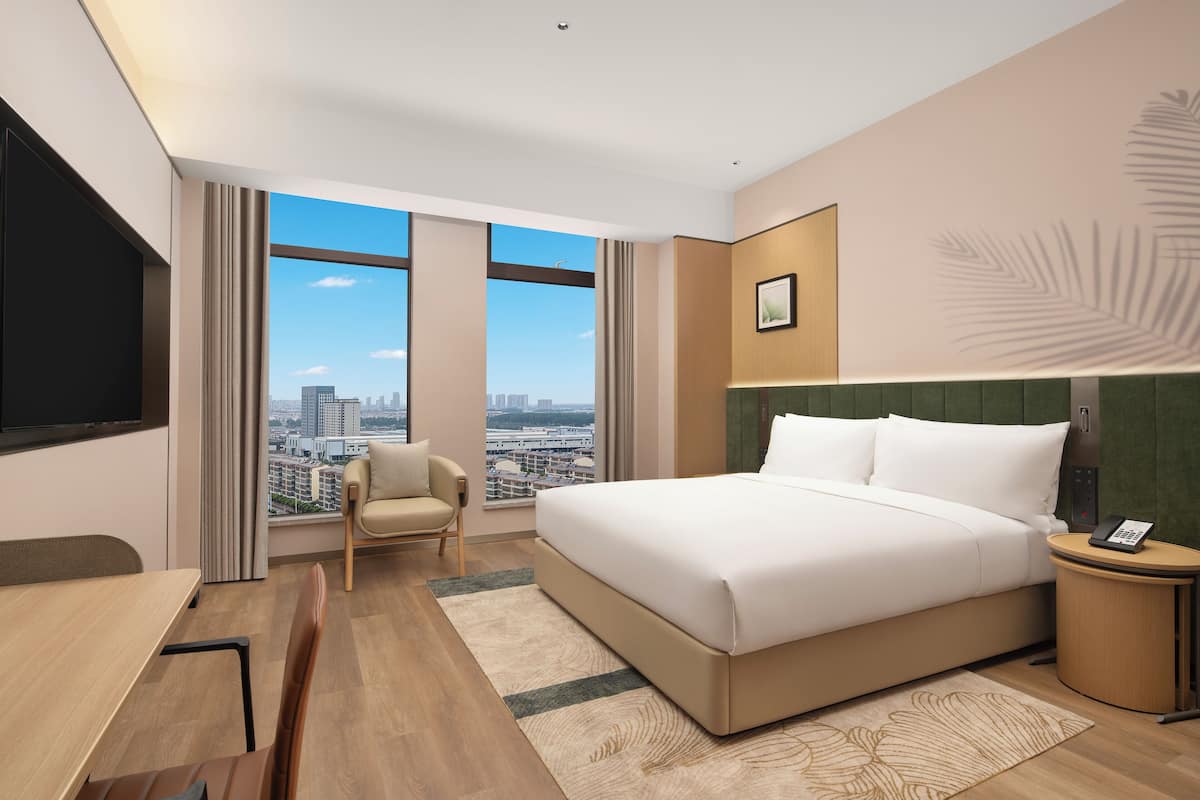 A hotel room with a large bed, a desk, a chair, a small table, and a view of the city through large windows.