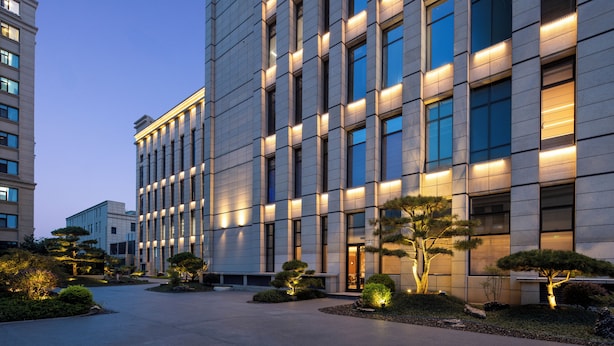 Un edificio moderno con fachada simétrica, amplios ventanales y un área ajardinada bien cuidada con árboles y arbustos.