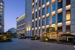 Un edificio moderno con fachada simétrica, amplios ventanales y un área ajardinada bien cuidada con árboles y arbustos.