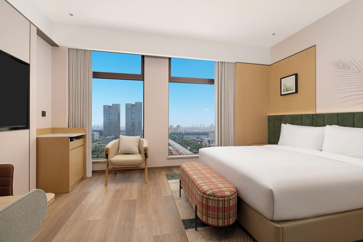A hotel room with a large bed, a chair, a desk, and a view of the cityscape through large windows.