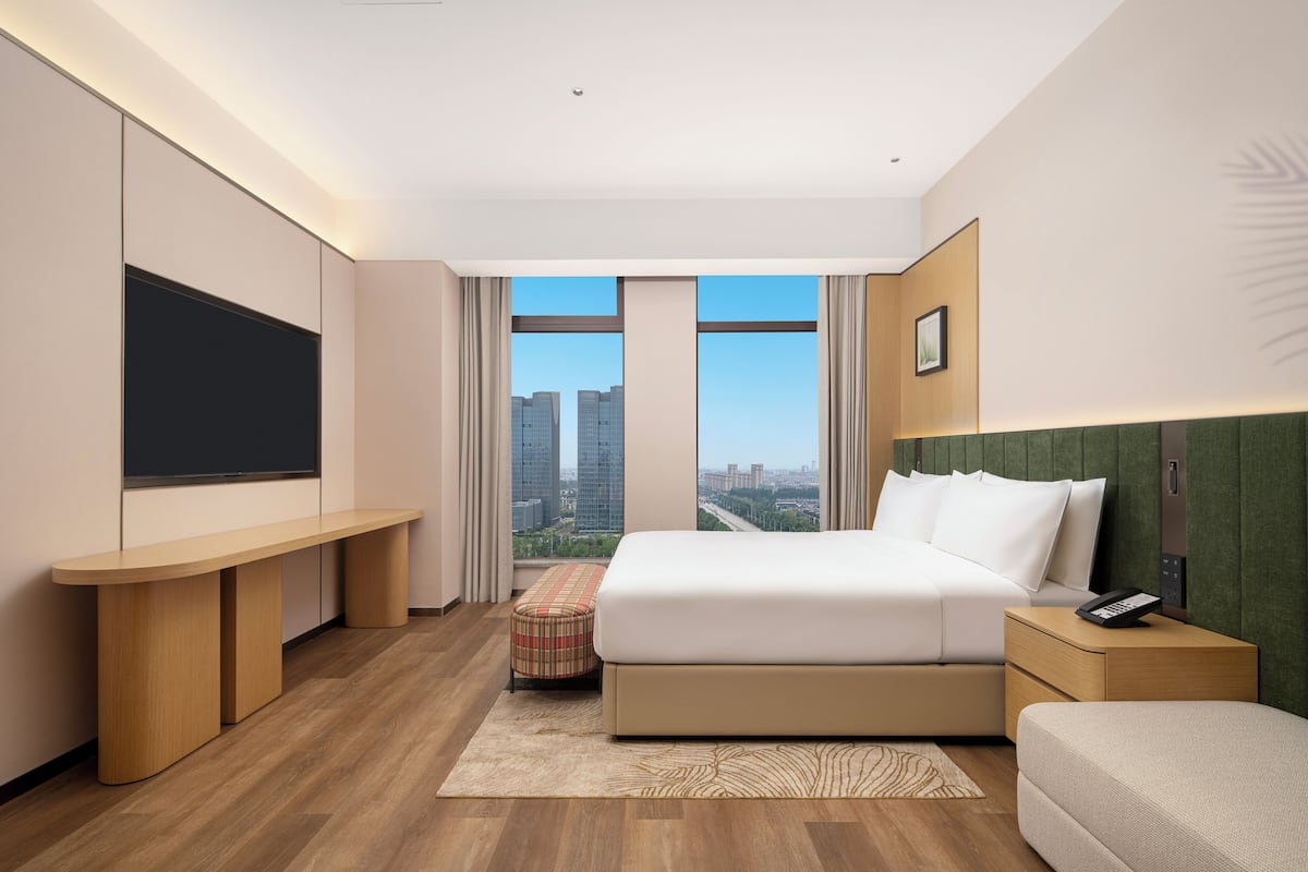 A modern hotel room with a large bed, a flat-screen TV, a desk, and a view of the city.