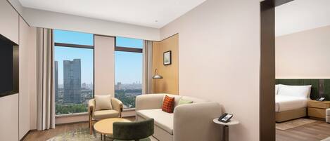 A modern hotel room with a large window, a sofa, a chair, a small table, a bed, and a bedside table with a phone.