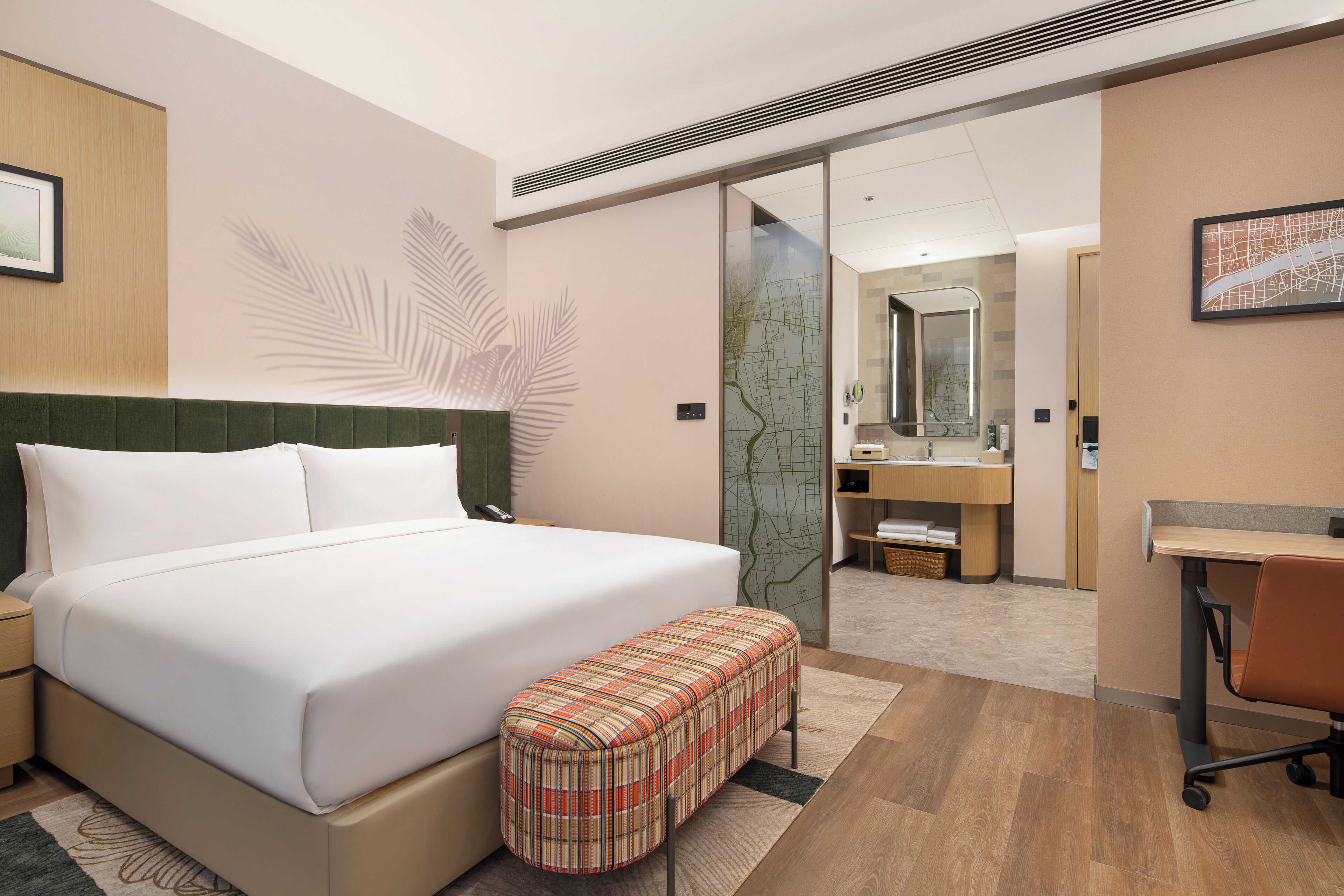 A hotel room with a large bed, a bench, a desk, and a bathroom visible through a glass door.