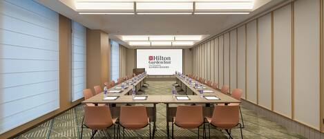 Un salón de conferencias con una larga mesa, sillas y una pantalla proyectora que muestra el logotipo de Hilton Garden Inn.