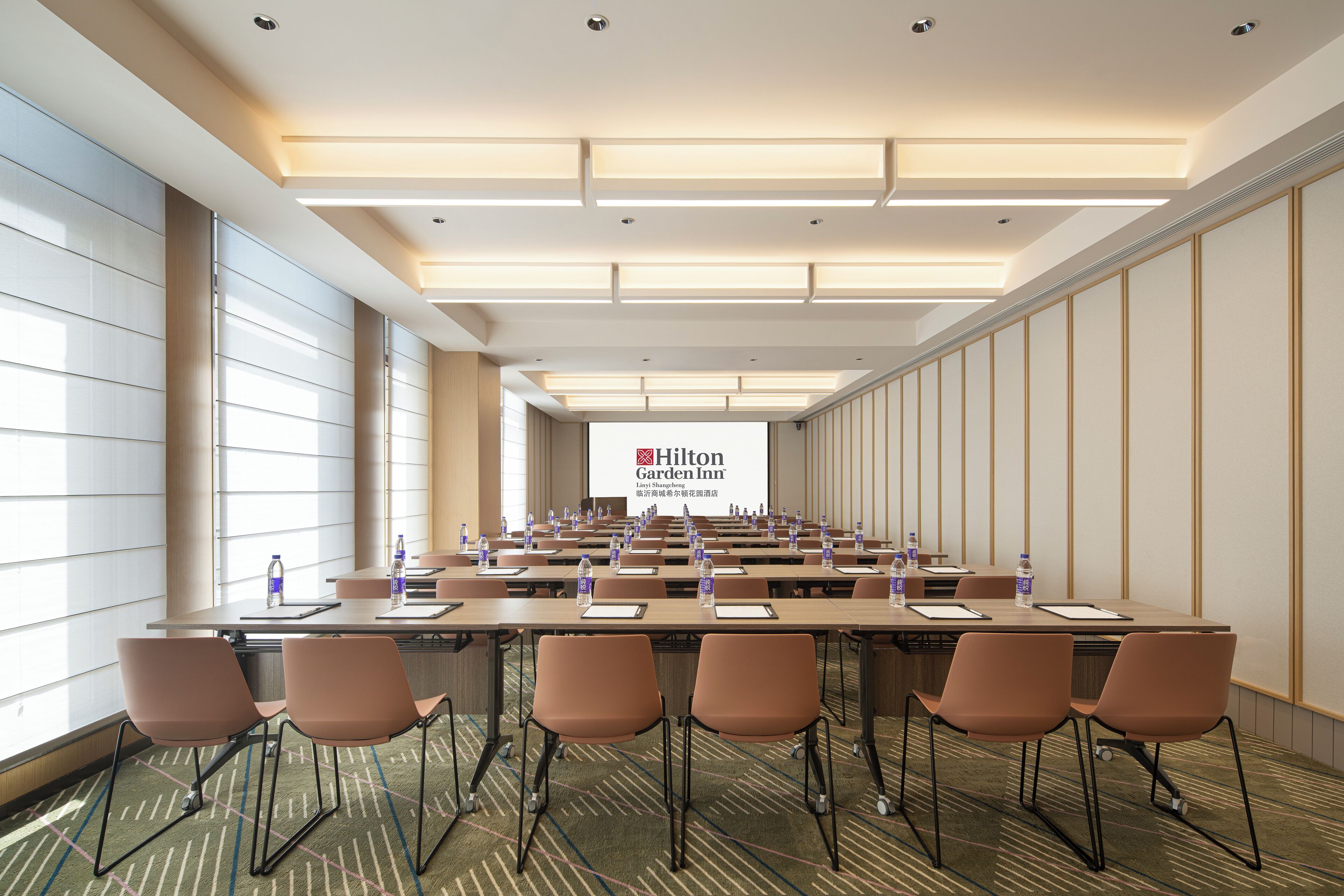 Una sala conferenze con un lungo tavolo, sedie, bottiglie d'acqua e uno schermo che mostra il logo Hilton Garden Inn.