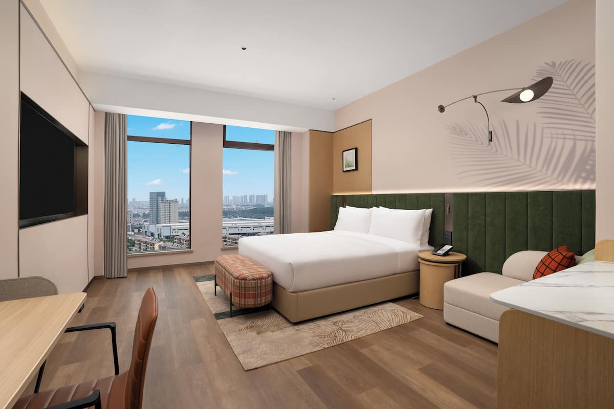 A modern hotel room with a large bed, a desk, a chair, a small sofa, and a view of the city through large windows.