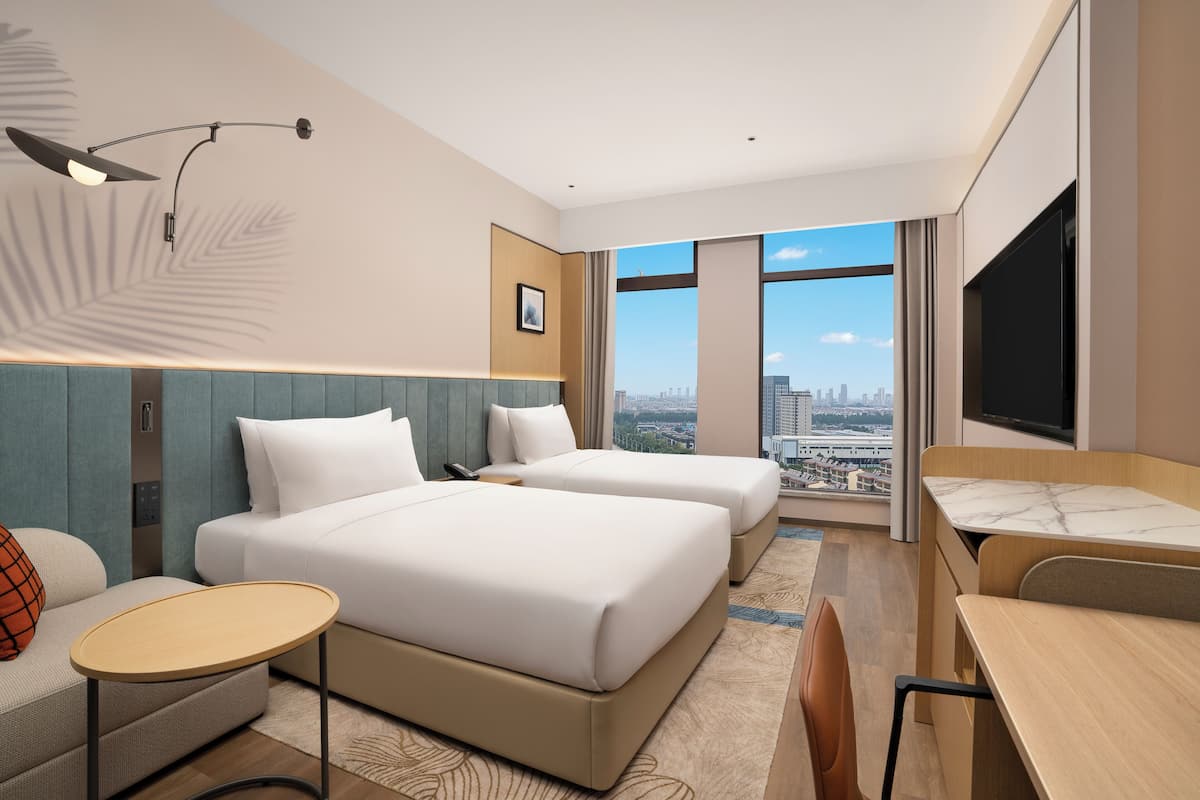 A hotel room with two beds, a desk, a chair, a small table, a TV, and a city view.
