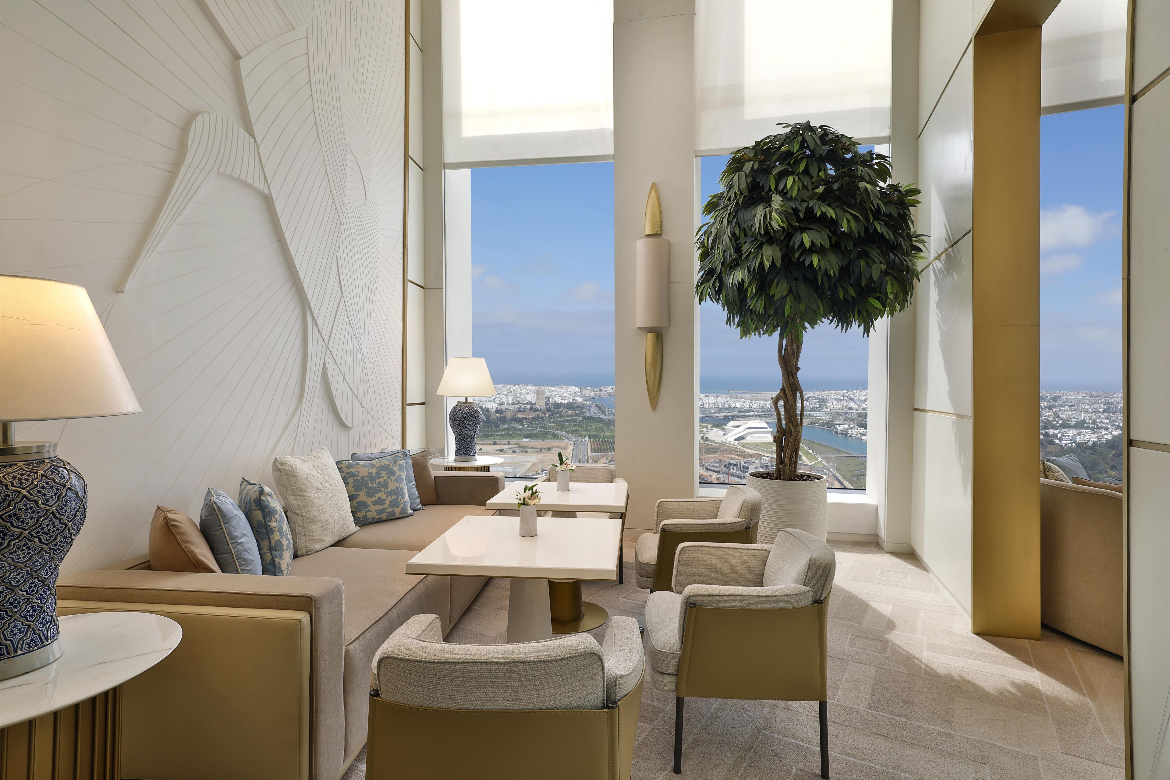 Une chambre d’hôtel moderne dotée d’une grande fenêtre donnant sur la ville, d’une plante en pot et d’un coin salon avec une table et des chaises.