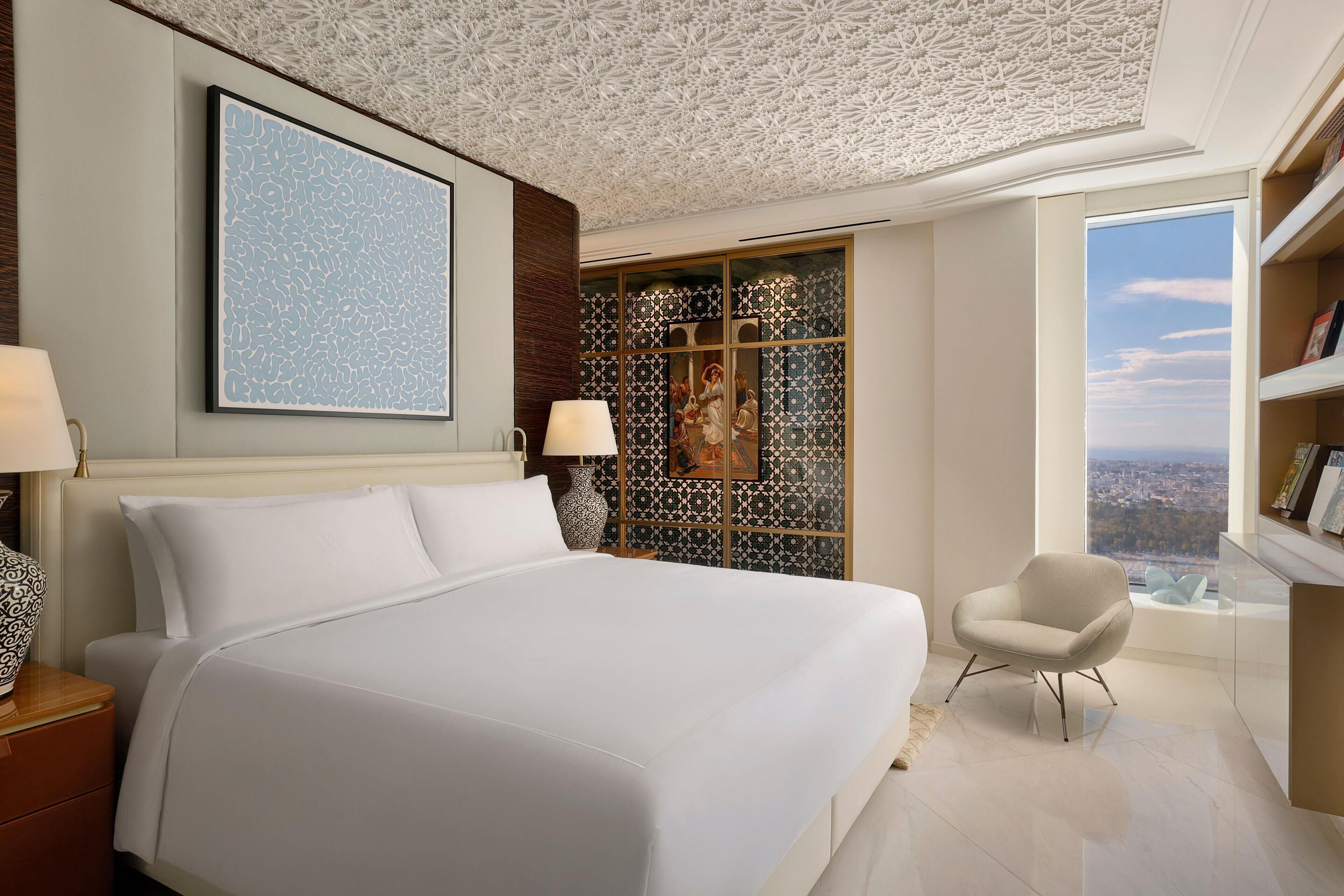 Habitación de hotel con una cama grande, una silla, vistas de la ciudad y un detalle decorativo en la pared.