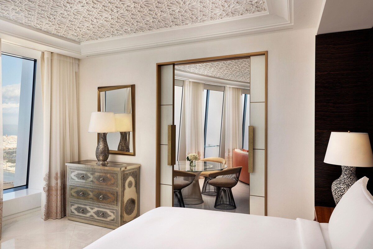 Habitación de hotel con cama, escritorio con lámpara, silla, mesa de comedor con sillas y vistas a través de una puerta.
