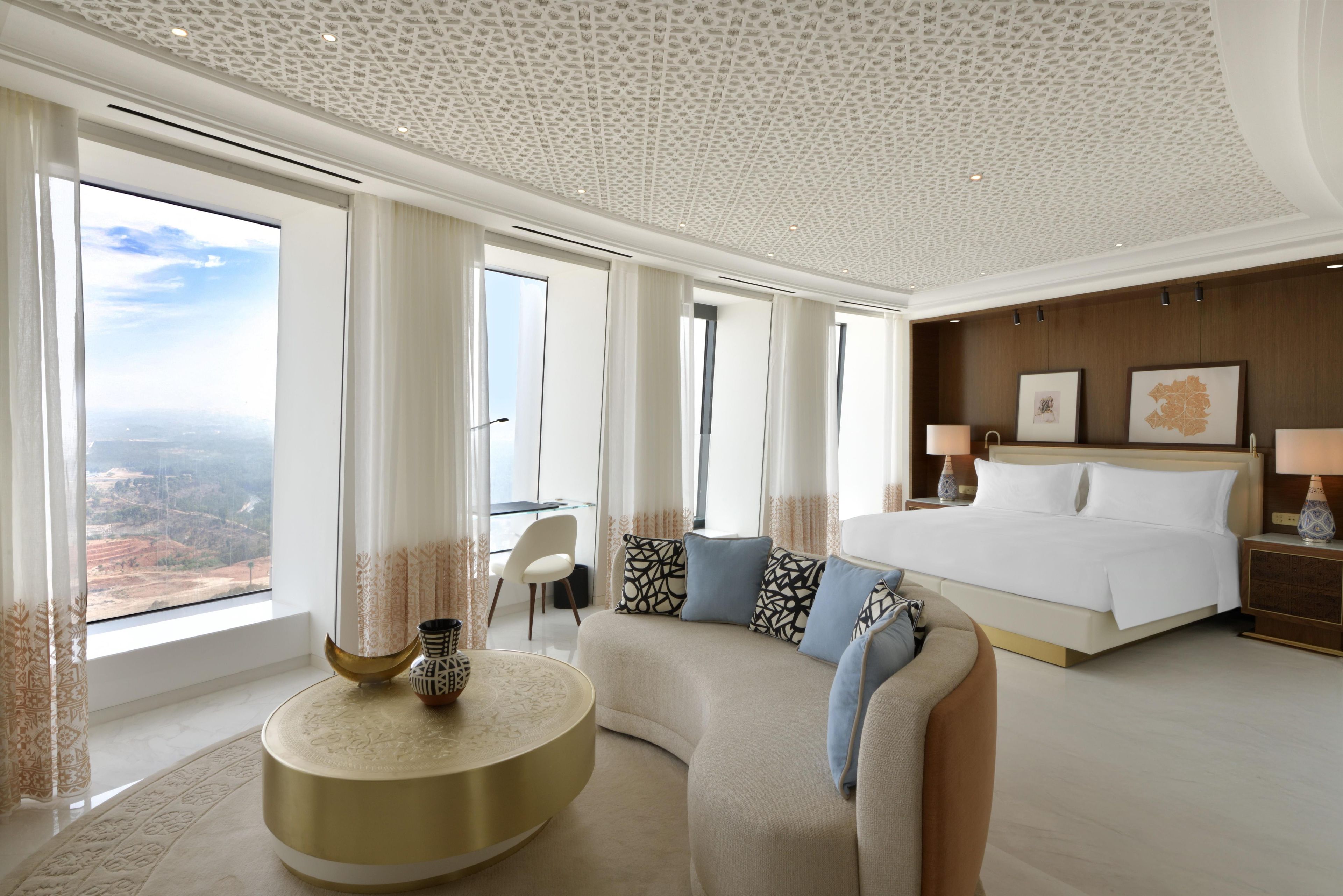 A modern hotel room with a large bed, a curved sofa, and a view of the landscape through large windows.