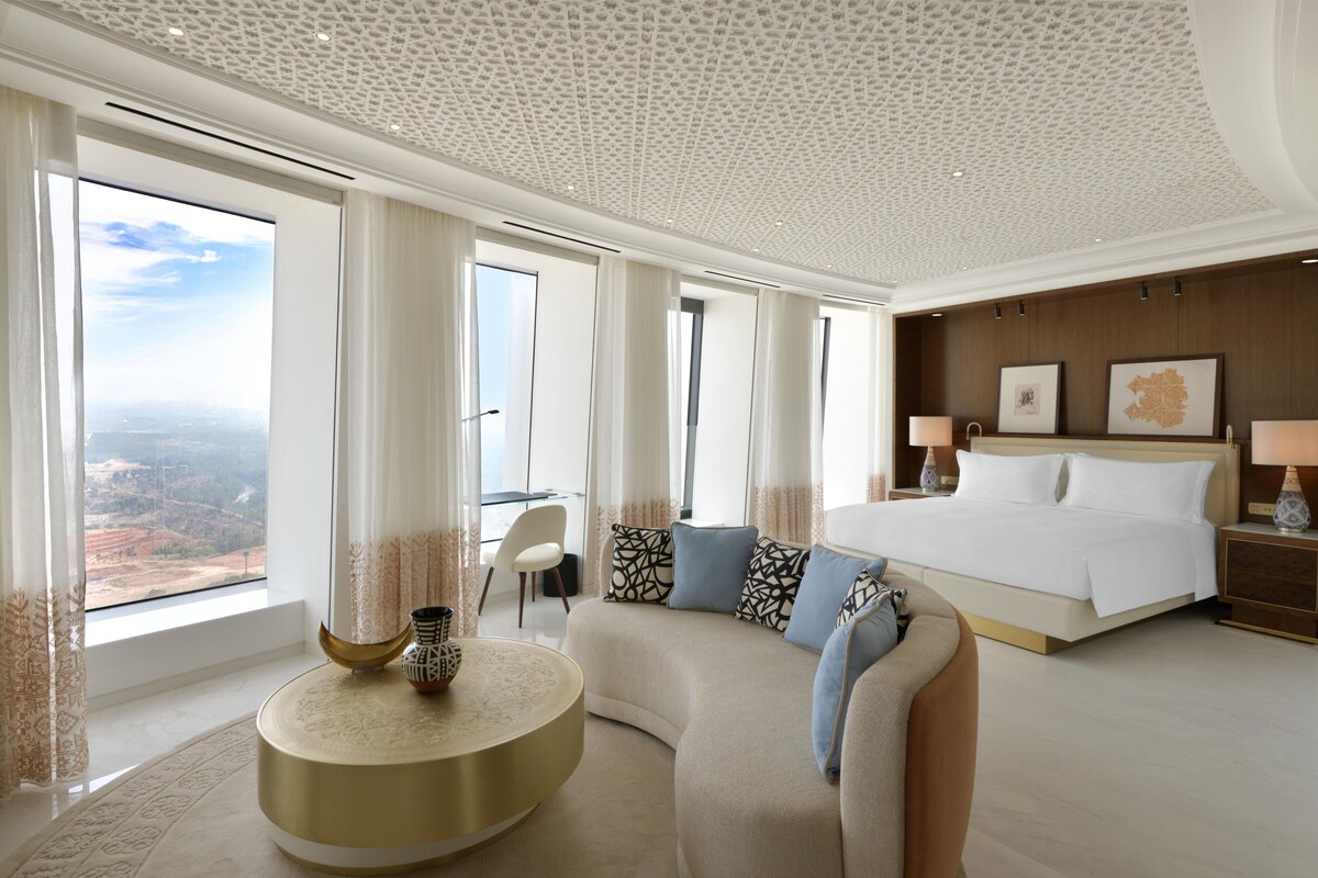 A modern hotel room with a large bed, a curved sofa, a round coffee table, and a view of the landscape through large windows.