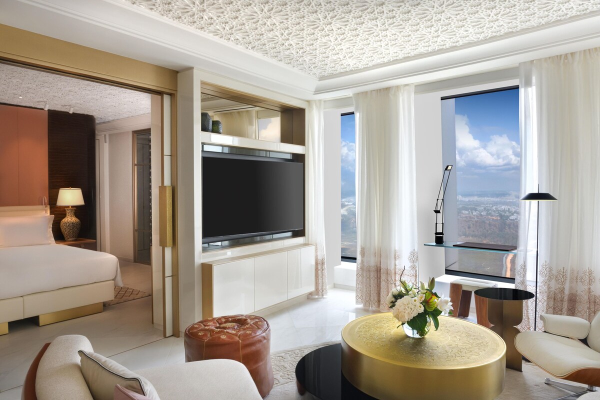 Una habitación de hotel moderna con una cama grande, un televisor de pantalla plana, un área de comedor con una mesa redonda y vistas de la ciudad a través de amplios ventanales.