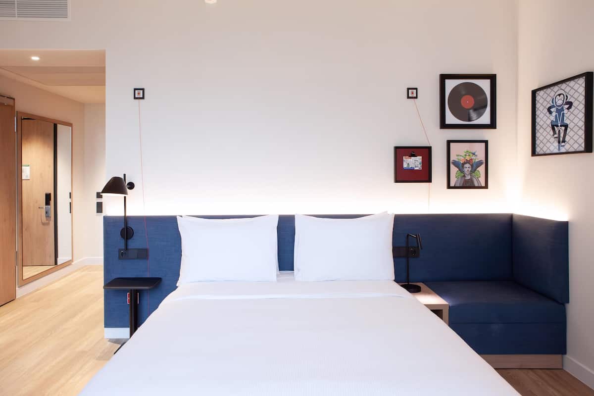 A modern hotel room with a large bed, a blue sofa, a bedside table, and a wall with framed artwork.