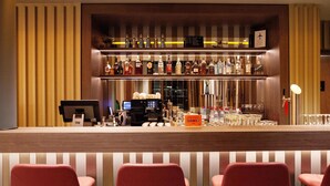 Angolo bar con un bancone in legno, diverse bottiglie di liquori, una cassa e tre sedie rosse.