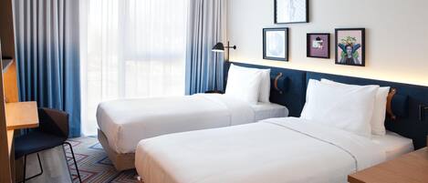 A hotel room with two beds, a desk, a chair, and a large window with curtains.