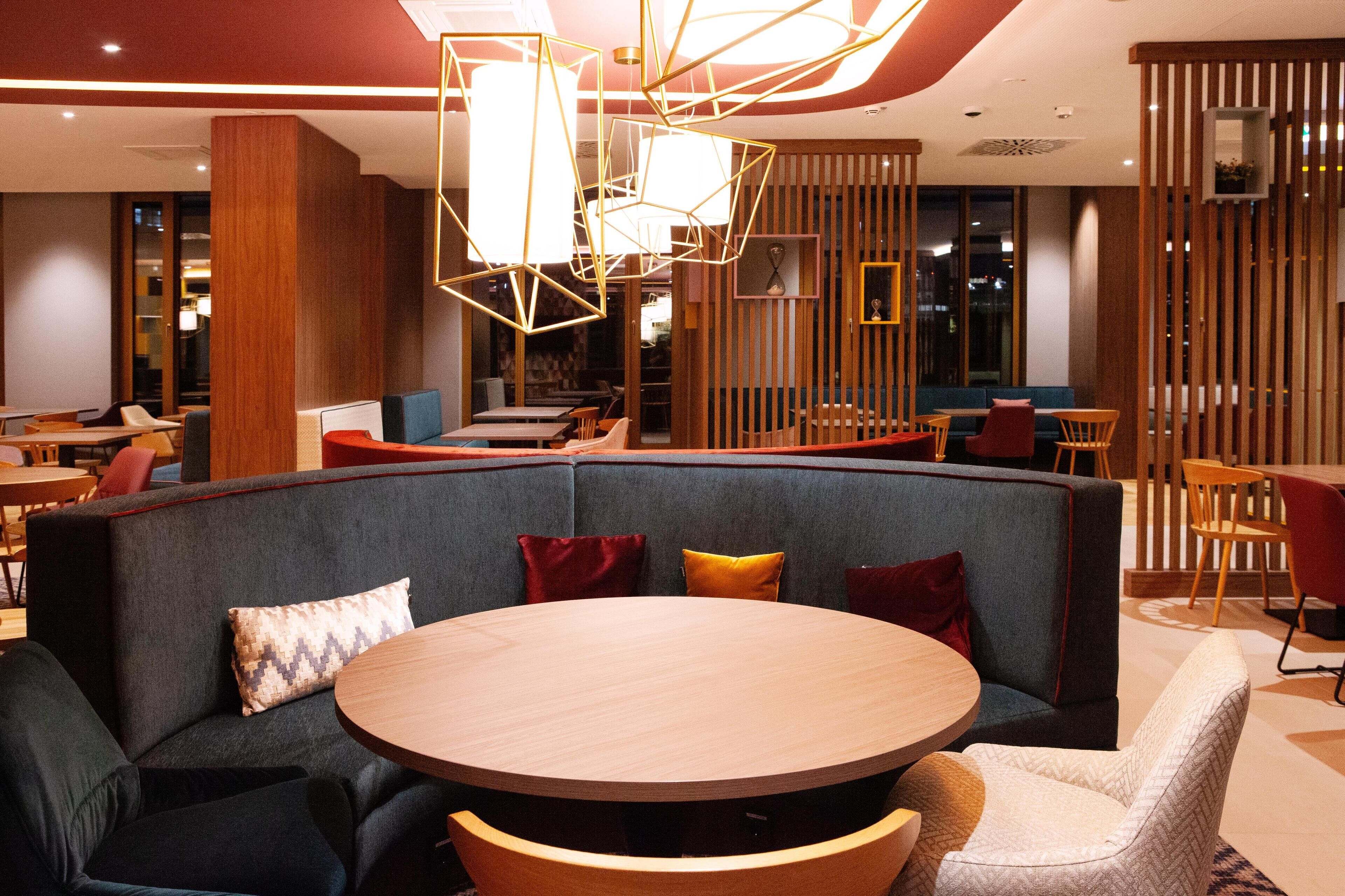 A modern hotel lobby with a round wooden table, cushioned seating, and geometric lighting fixtures.