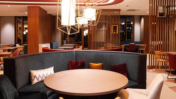 A modern hotel lobby with a round wooden table, cushioned seating, and geometric lighting fixtures.