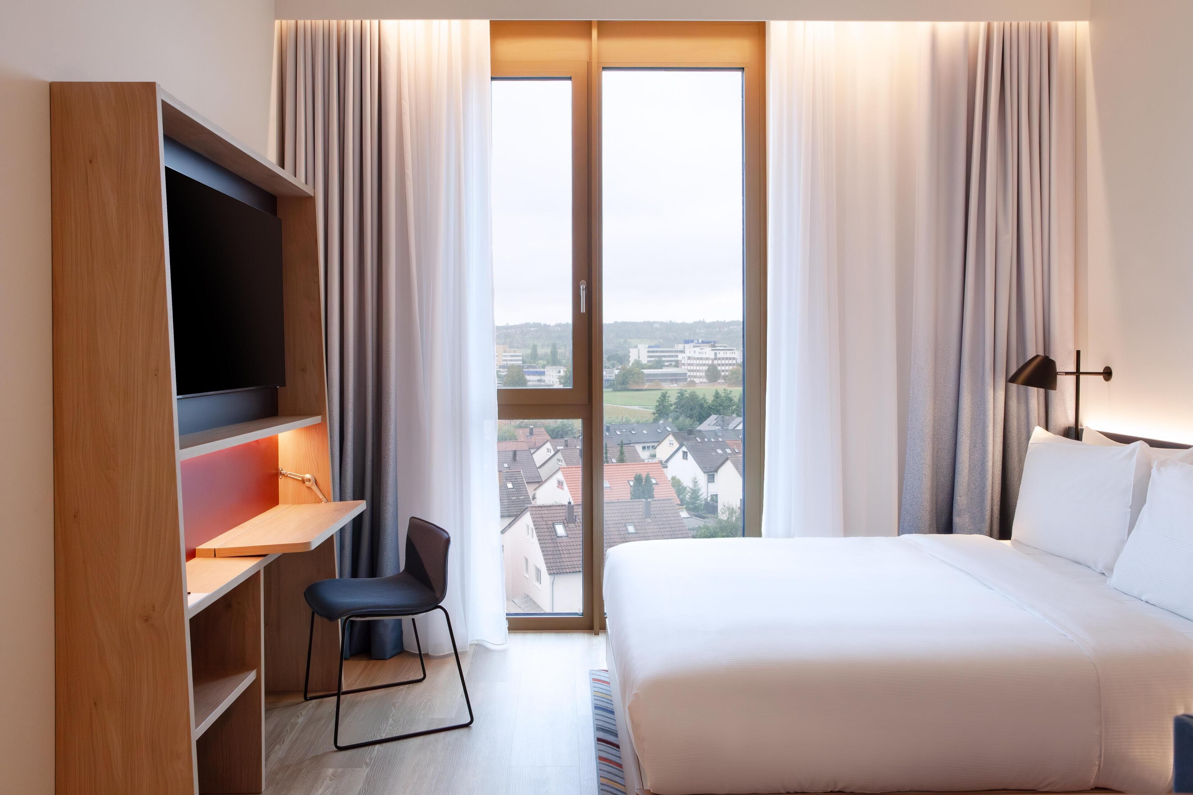 Habitación de hotel con cama, escritorio con silla, televisor y ventana grande con cortinas.