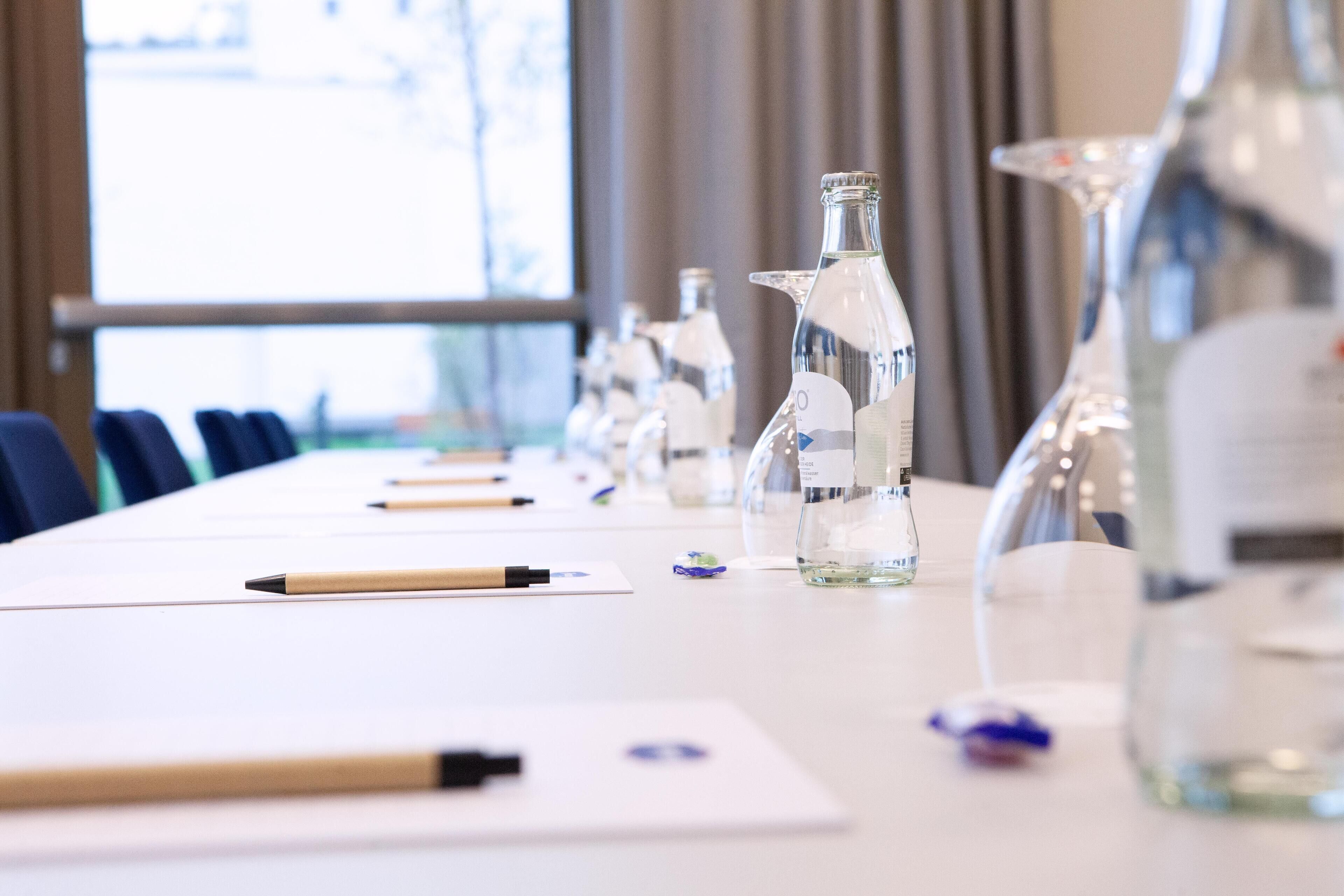 A conference table with glassware and pens, set in a room with a large window and curtains.