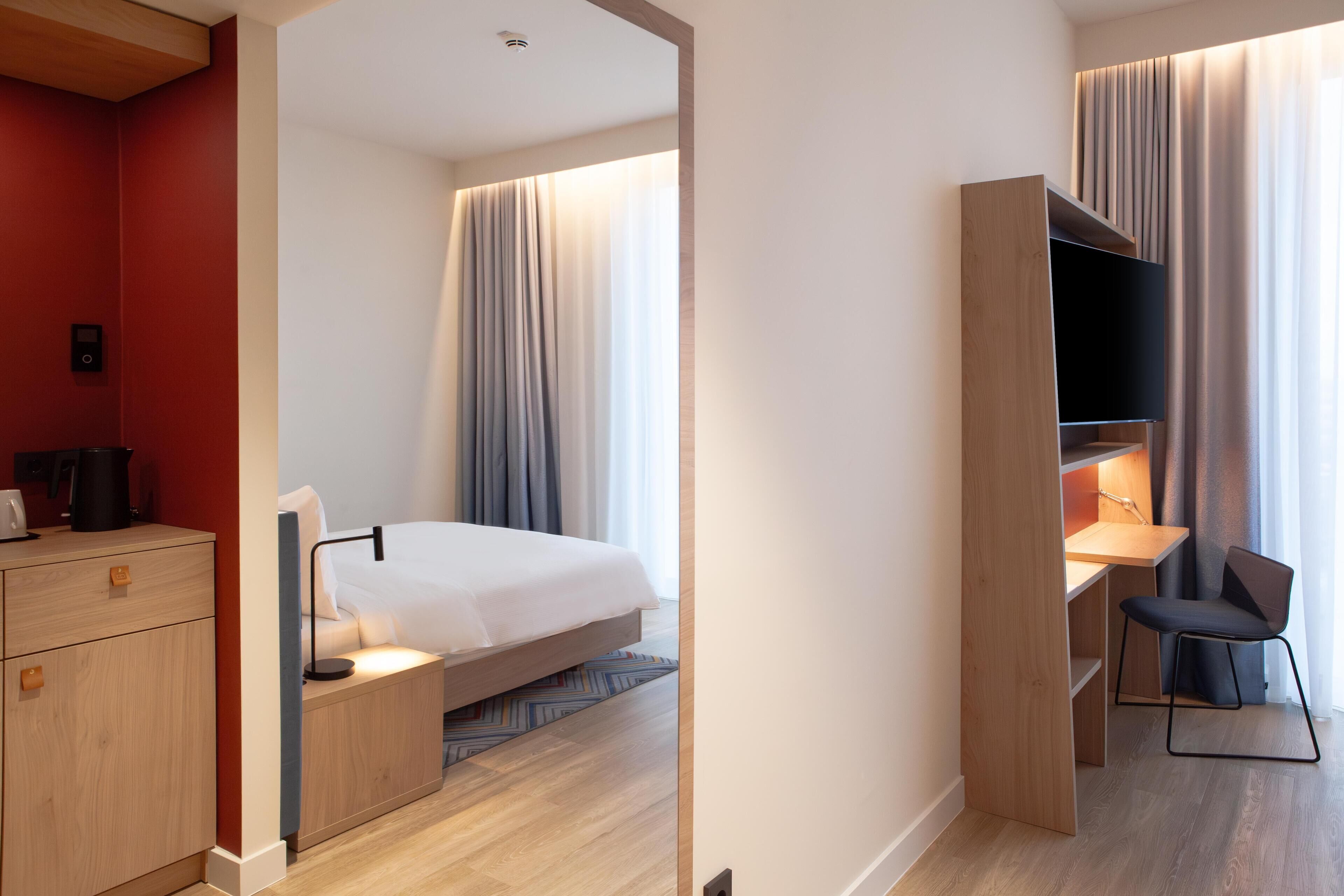 A modern hotel room with a bed, bedside table, desk, chair, and a wardrobe.