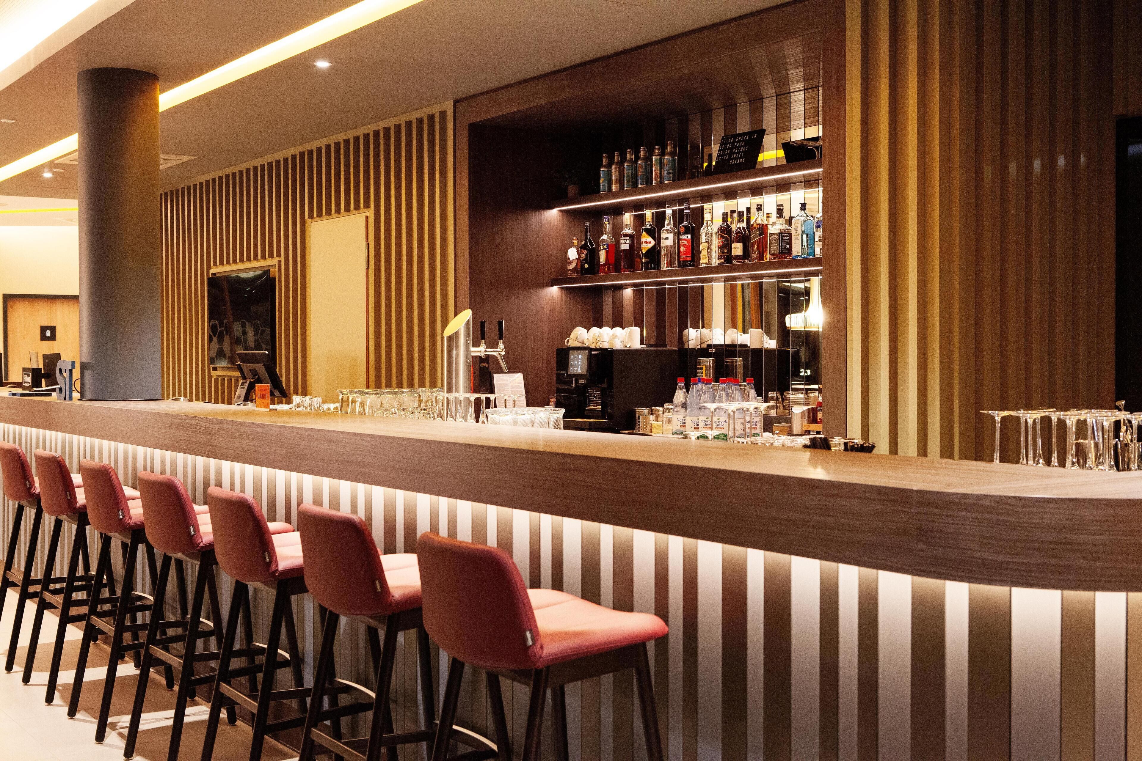 A bar area with a long counter, multiple stools, and shelves stocked with bottles and glasses.