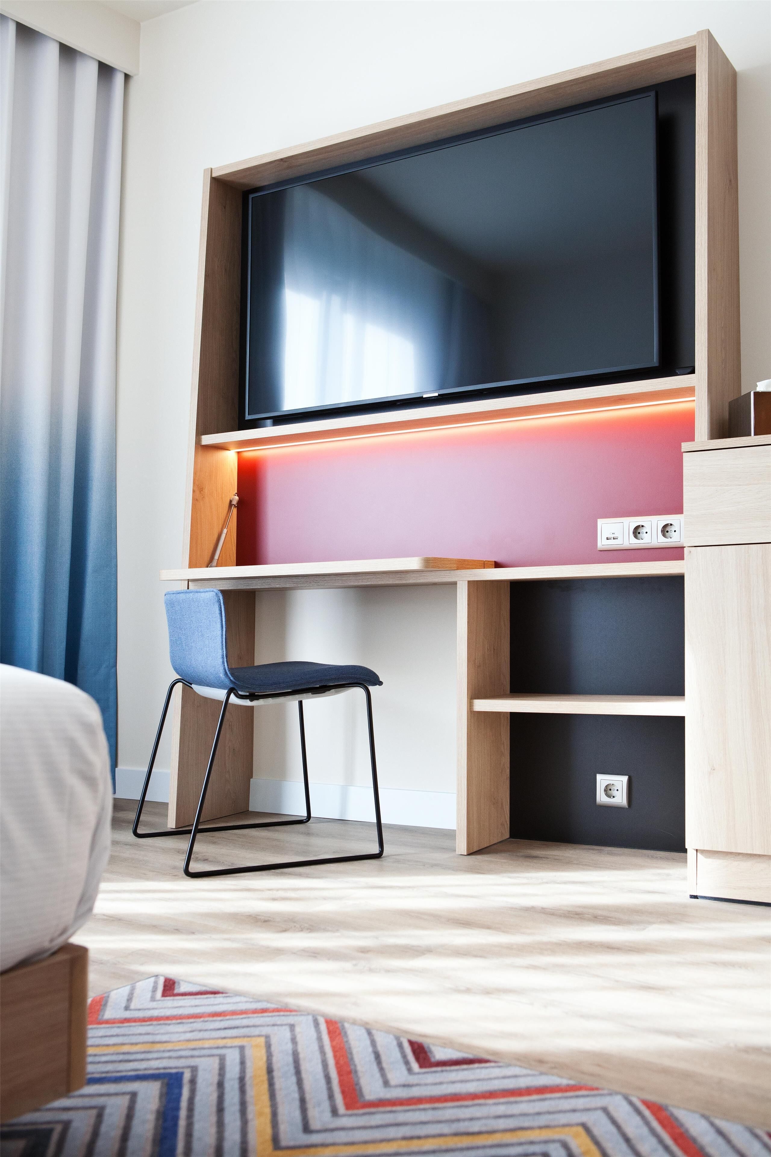 A modern hotel room with a large flat-screen TV mounted on a wooden wall, a blue chair, and a colorful geometric-patterned rug.