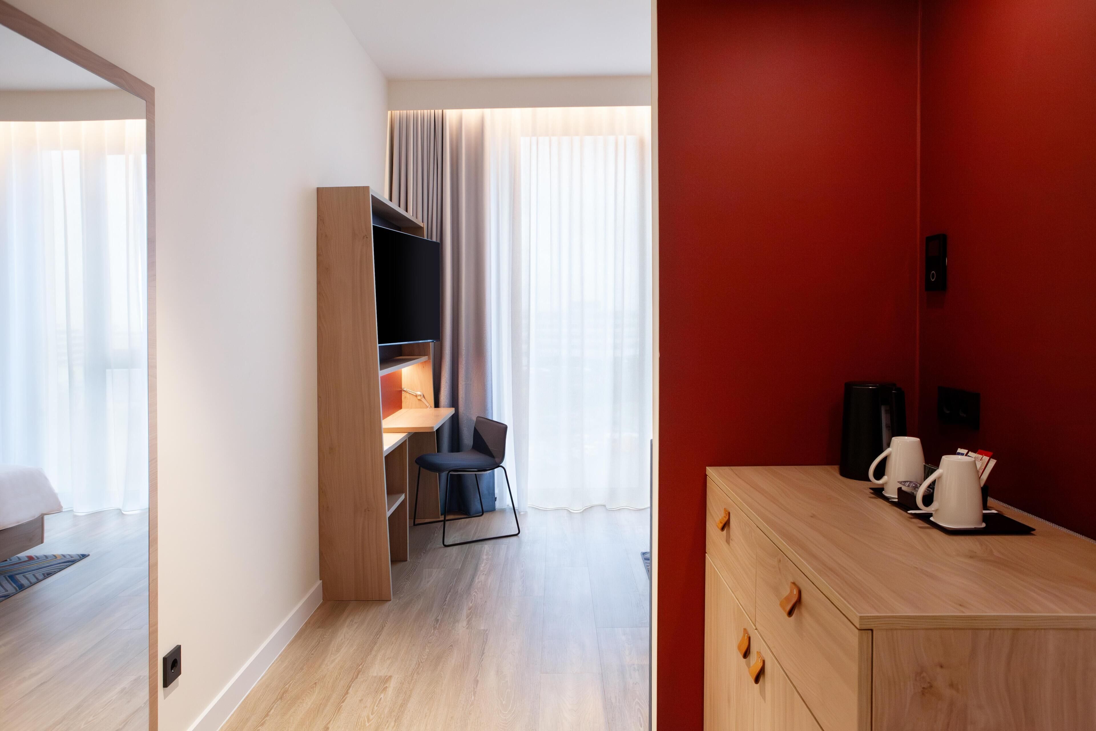 A hotel room with a wooden desk, a chair, a wardrobe, and a coffee station.