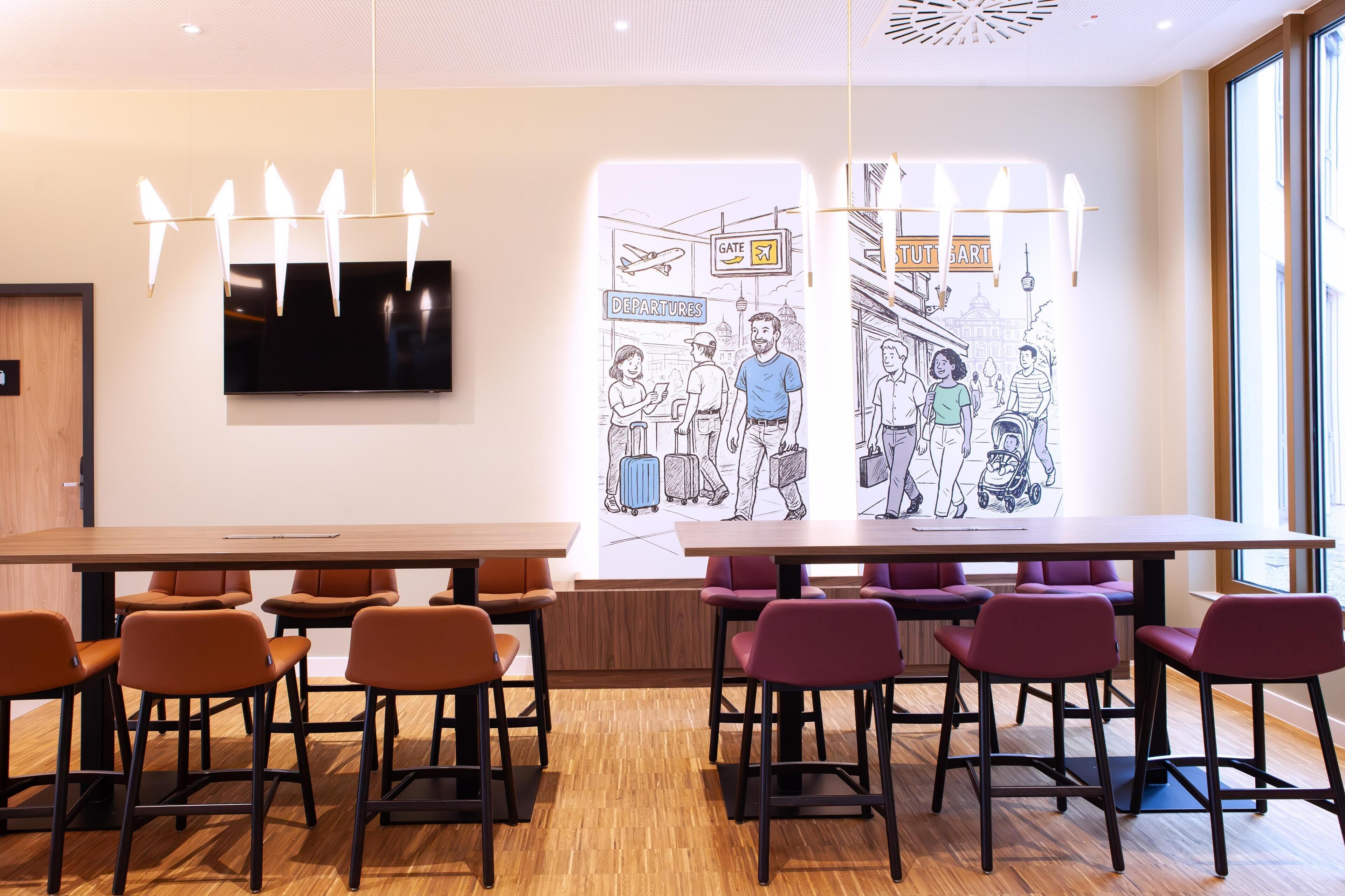 A modern hotel lobby with a long wooden table, several orange chairs, and a large wall mural depicting a city scene.
