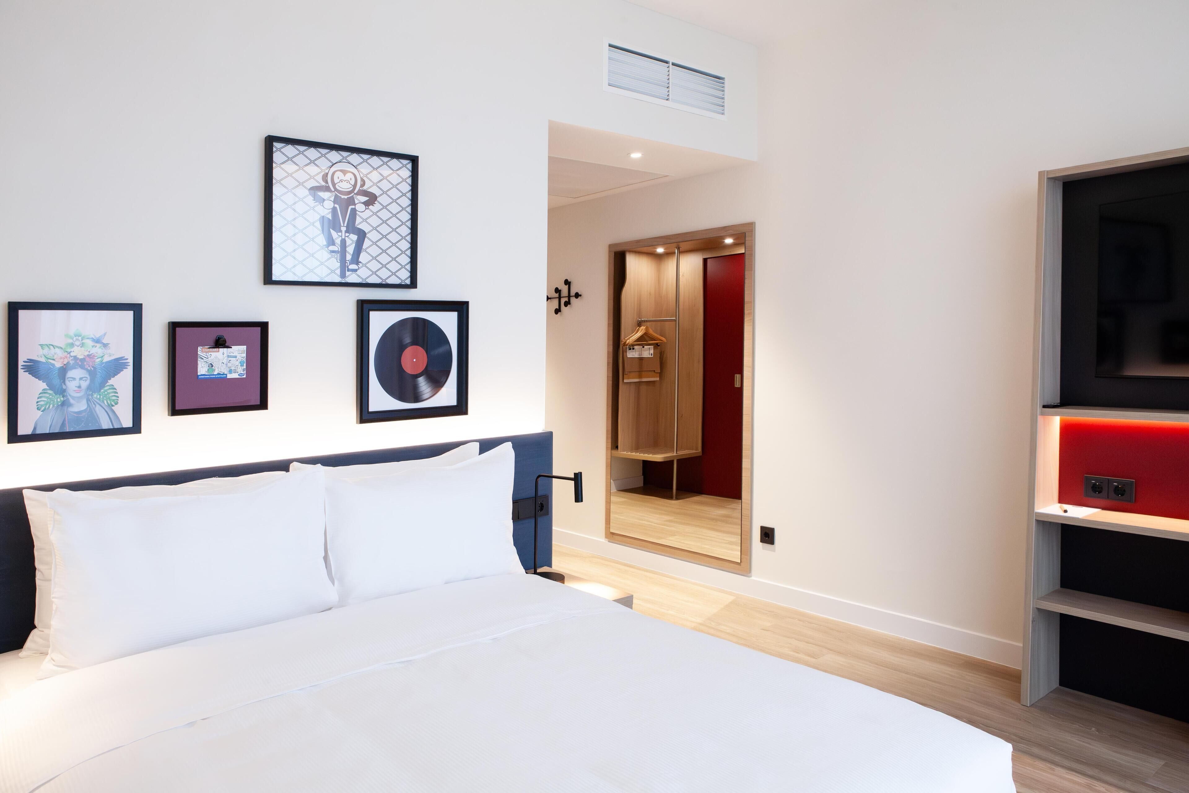A modern hotel room with a large bed, a wardrobe, a mirror, and framed artwork on the wall.