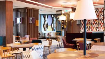 A modern hotel lobby with wooden furniture, a large lamp, and geometric wall patterns.