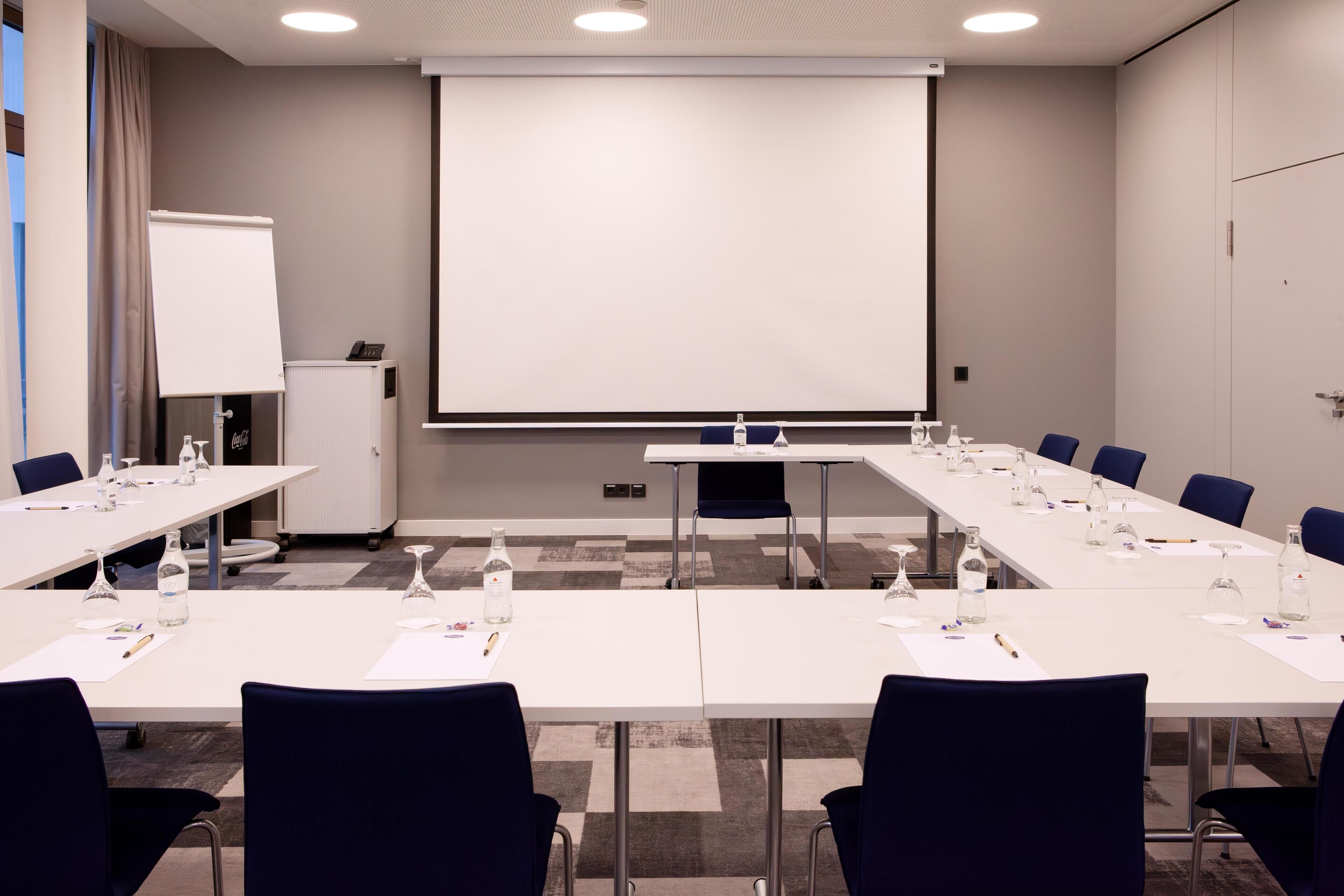 A conference room with long tables, chairs, a projector screen, and a whiteboard.