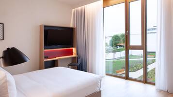 A hotel room with a large bed, a flat-screen TV, a desk with a red accent, and a view of a park through large sliding doors.
