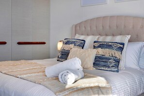 Ein ordentlich bezogenes Bett mit einer gestreiften Decke, zwei Kissen mit blau-weißem Muster und einem aufgerollten Handtuch.