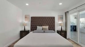 Un dormitorio moderno con cabecera capitonada, una cama grande, dos mesitas de noche con lámparas y vistas a través de una puerta corrediza de vidrio.