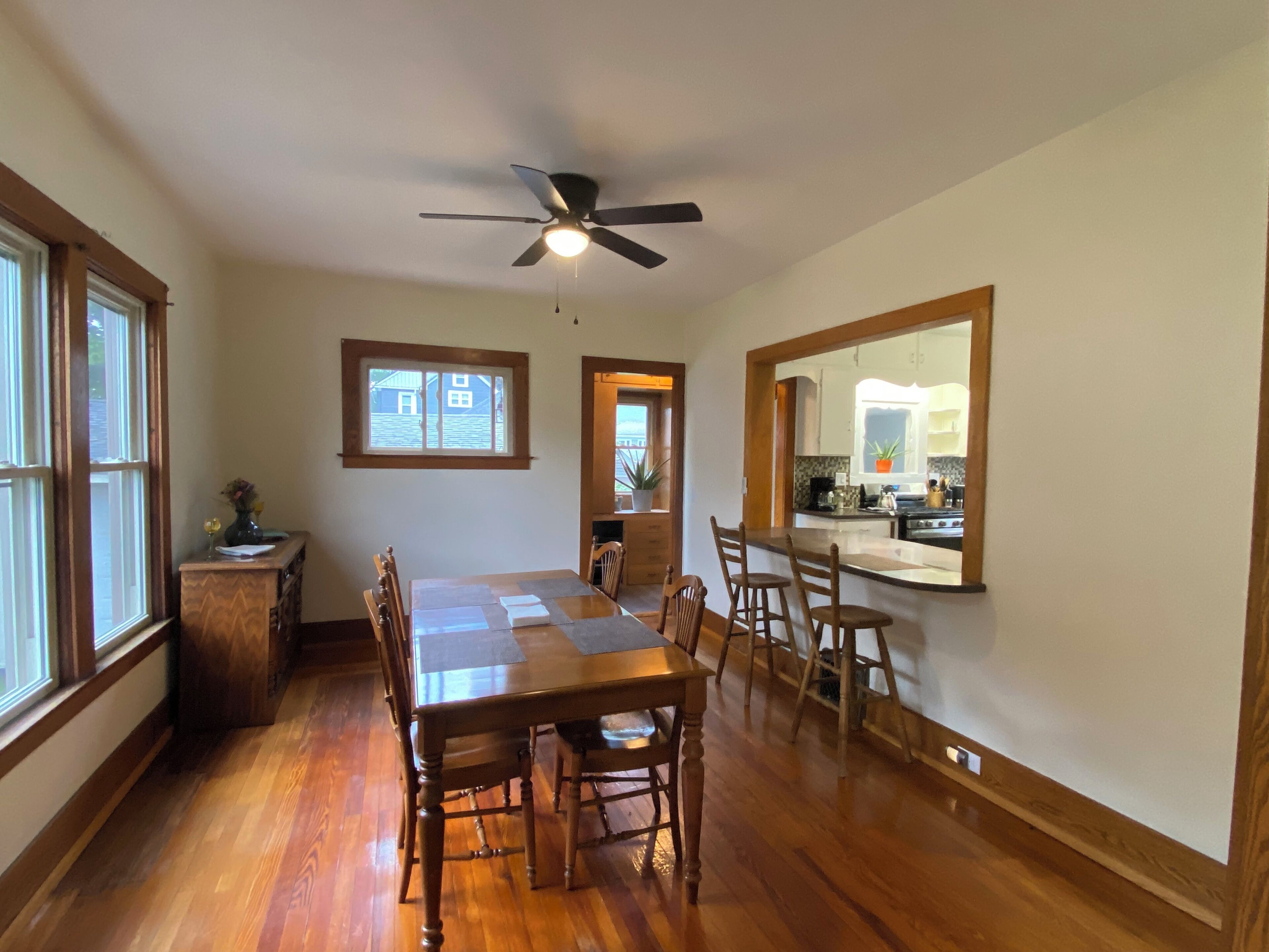 Un coin repas avec une table et des chaises en bois, un ventilateur de plafond et une vue sur la cuisine grâce à un miroir.