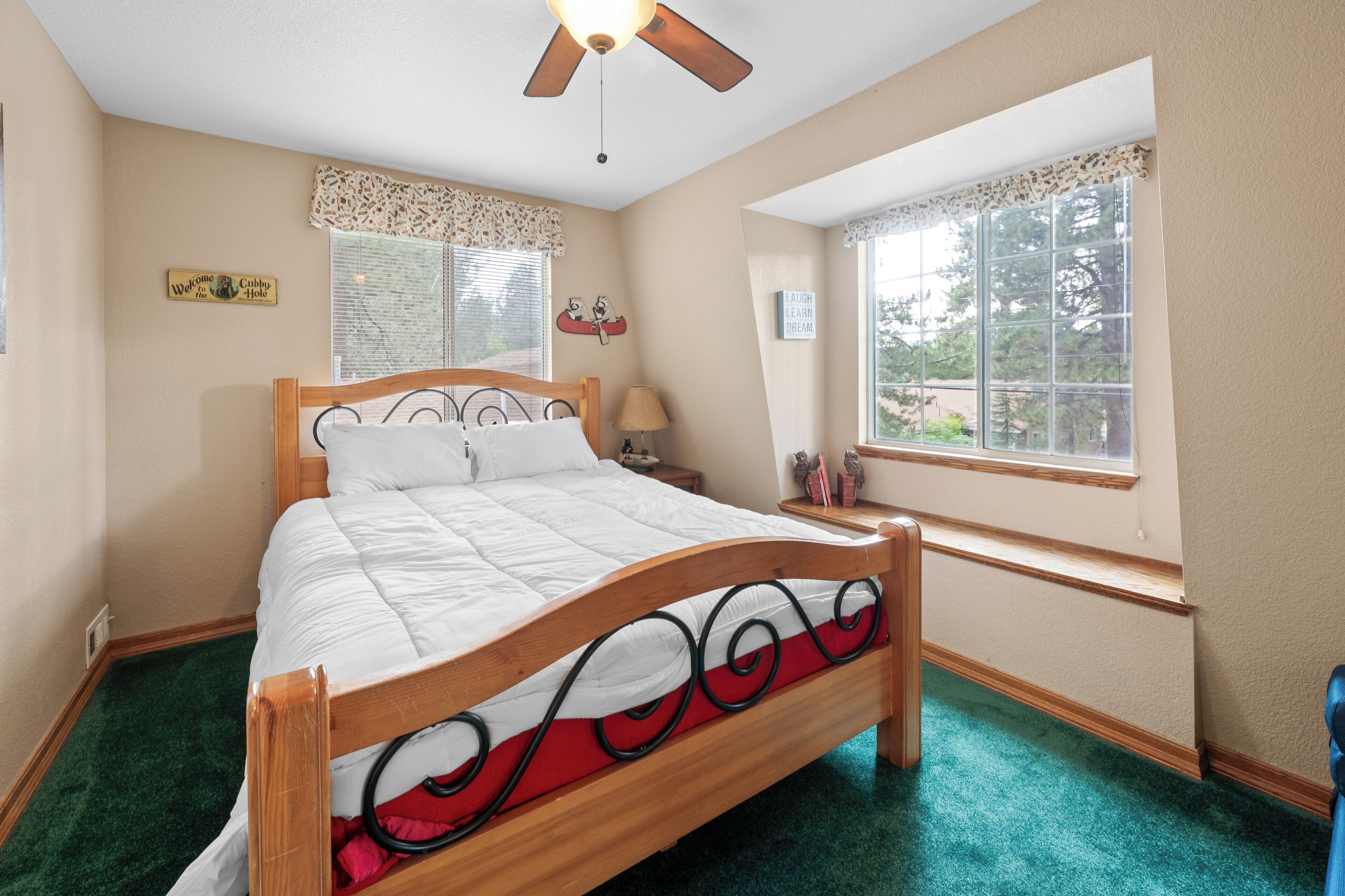 A bedroom with a wooden bed, a ceiling fan, a window with a view, and a green carpet.
