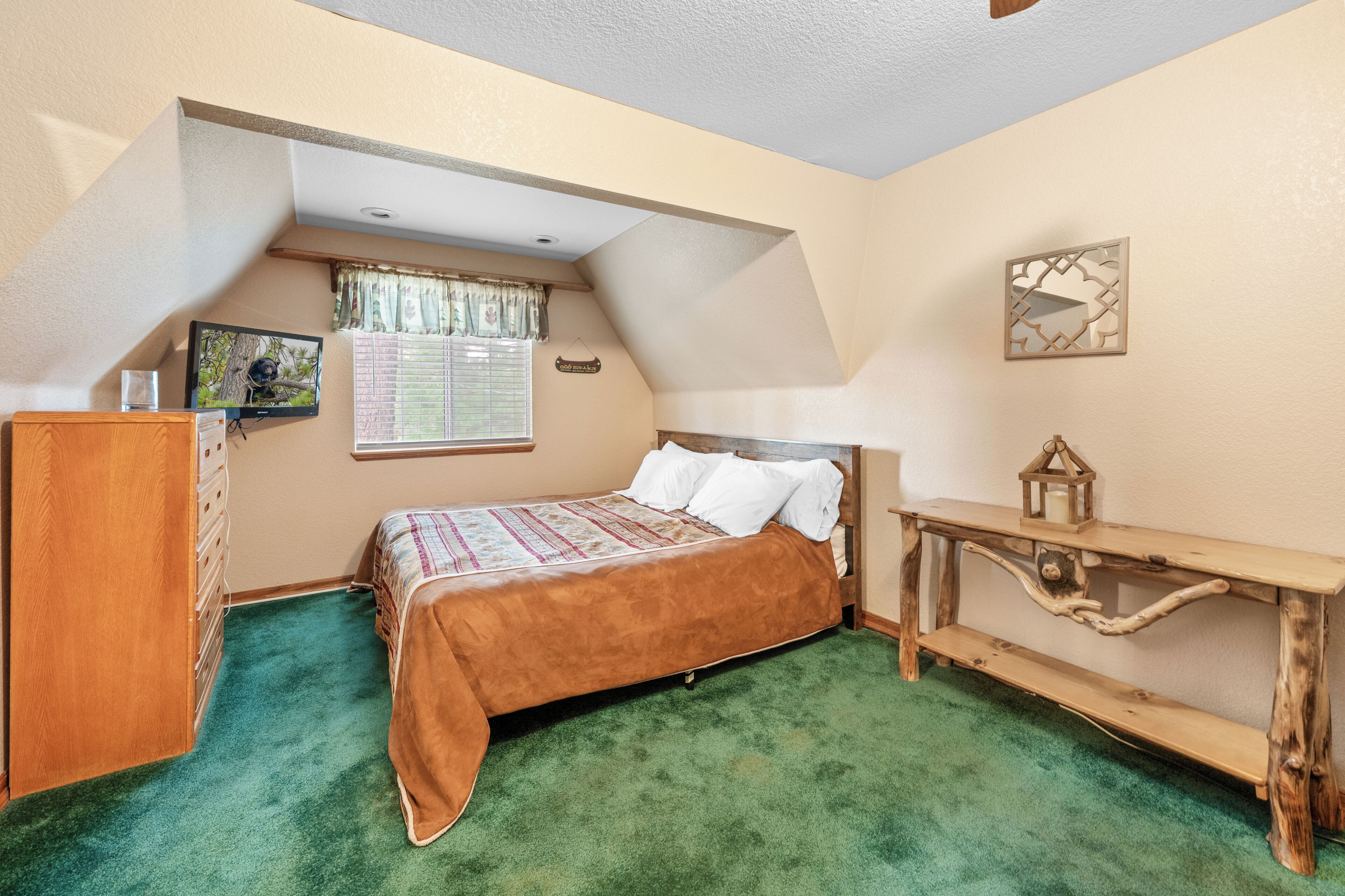 Ein Schlafzimmer mit schrägen Wänden, einem Bett mit grünem Teppich, einer Holzkommode, einem Nachttisch und einem kleinen Tisch mit einer Laterne.