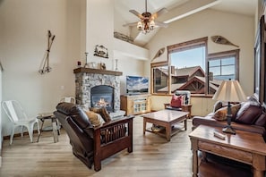 A spacious living room with a stone fireplace, wooden furniture, and a large window offering a view of the house.