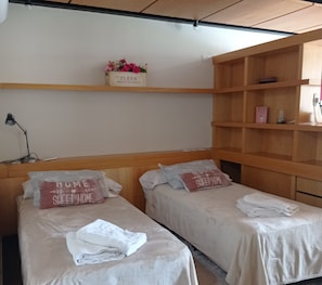 A hotel room with two single beds, each with a pillow and a folded towel. There is a shelf above the beds with a floral arrangement and a small box labeled "FLEUR PRODUIT DE FRANCE".