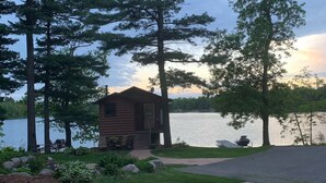 Une cabane dotée d’une terrasse donnant sur un lac, entourée de grands arbres et d’une pelouse bien entretenue.