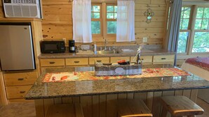 Une cuisine dotée d’un plan de travail en granit, de tabourets en bois, d’un réfrigérateur, d’un four à micro-ondes, d’un évier et d’une fenêtre avec des rideaux.