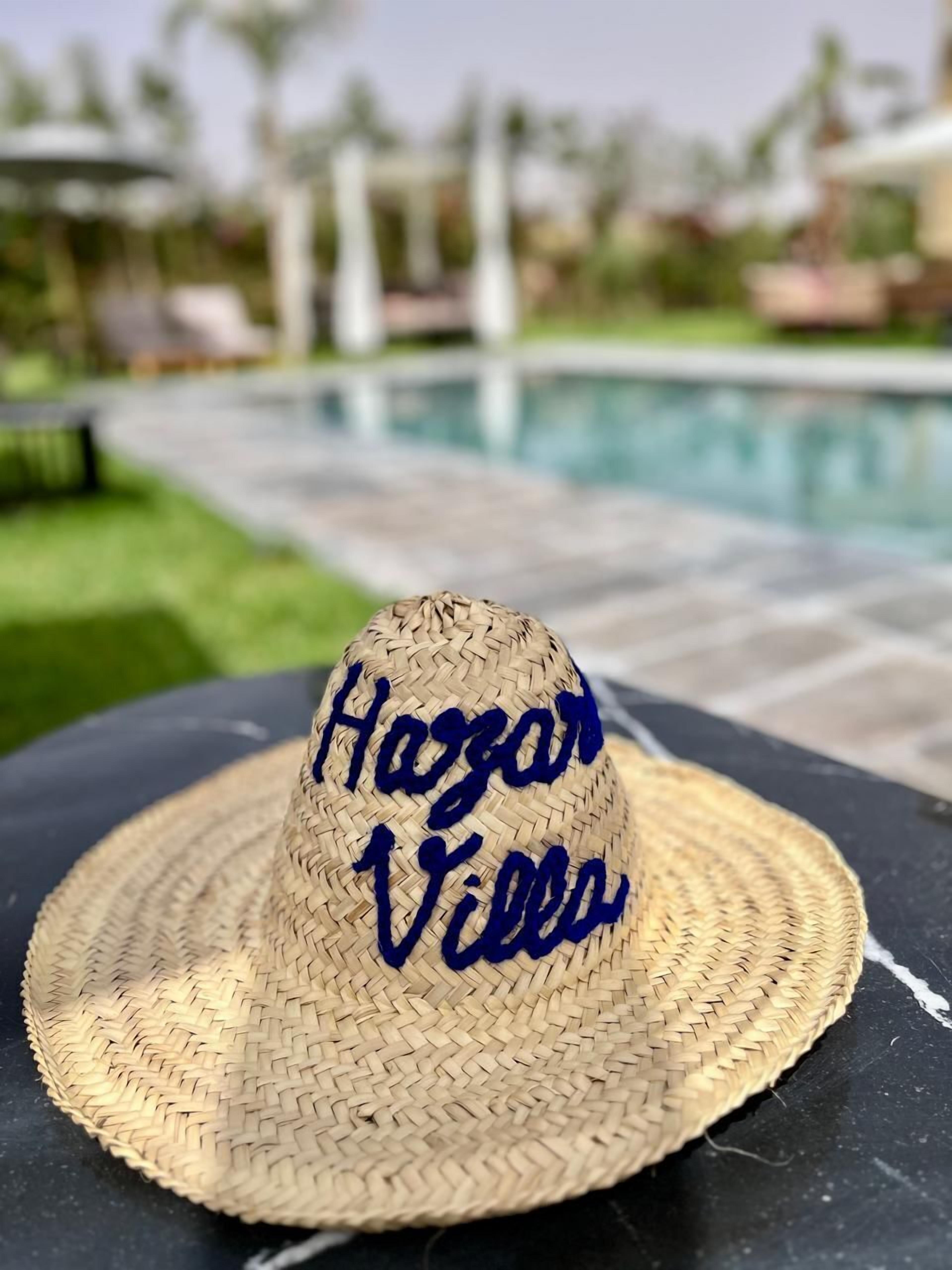 Ein Strohhut mit "Hazan Villes" Stickerei, auf einer schwarzen Oberfläche neben einem Pool platziert, mit unscharfem Hintergrund aus Grün und Gebäuden.