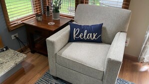 Un sillón gris claro con una almohada que dice 'Relax', un escritorio de madera con una planta y vistas a un balcón con vegetación.