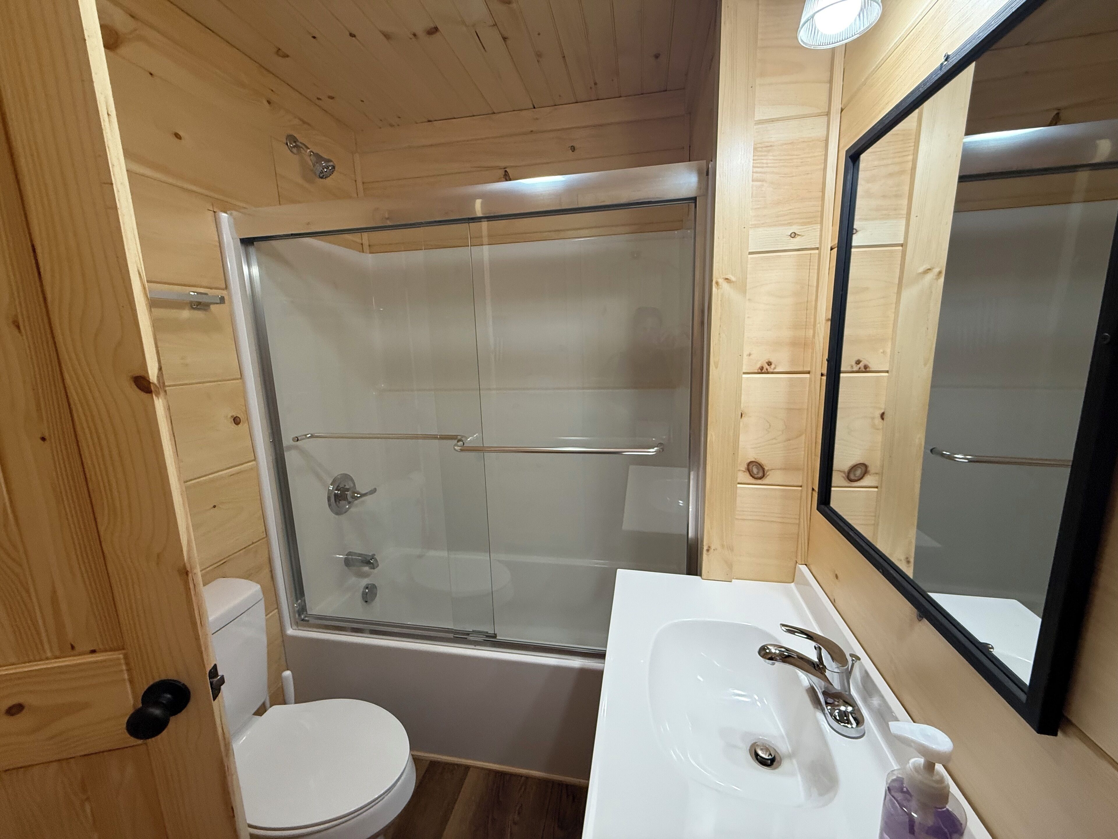 Une salle de bain rustique, avec des murs en bois, une douche en verre, des toilettes blanches et un lavabo agrémenté d’un miroir.